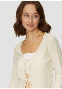 Cremfarbener, strukturierter Cardigan mit tiefem V-Ausschnitt und Bindegürtel. Darunter befindet sich ein weißes Tanktop. Schlichtes Design, ohne Muster.