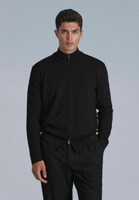 Veste zippée noire en matière tricotée avec un col haut, dotée d'une fermeture éclair à double sens à l'ourlet et de manches longues ajustées.