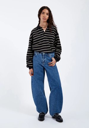 Frau mit lockigem Haar, die ein schwarz-grau gestreiftes langärmliges Shirt, weite blaue Jeans, schwarze Loafers und goldenen Schmuck trägt, steht vor weißem Hintergrund.