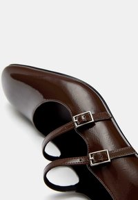 Pantof glossy de culoare maro închis, tip Mary Jane, cu două curele ajustabile cu catarame argintii și vârf rotunjit pe fundal alb.