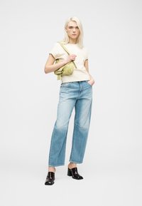 Ξανθιά γυναίκα με μπεζ μπλουζάκι, φαρδιά τζιν παντελόνια ανοιχτού μπλε, μαύρες mule με τακούνι και κρατώντας μια ανοιχτοπράσινη τσάντα ώμου.