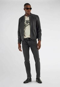 Sort læderjakke, cremefarvet grafisk t-shirt, grå jeans og sorte ankelstøvler. Model iført mørke solbriller, der står imod en ensfarvet baggrund.