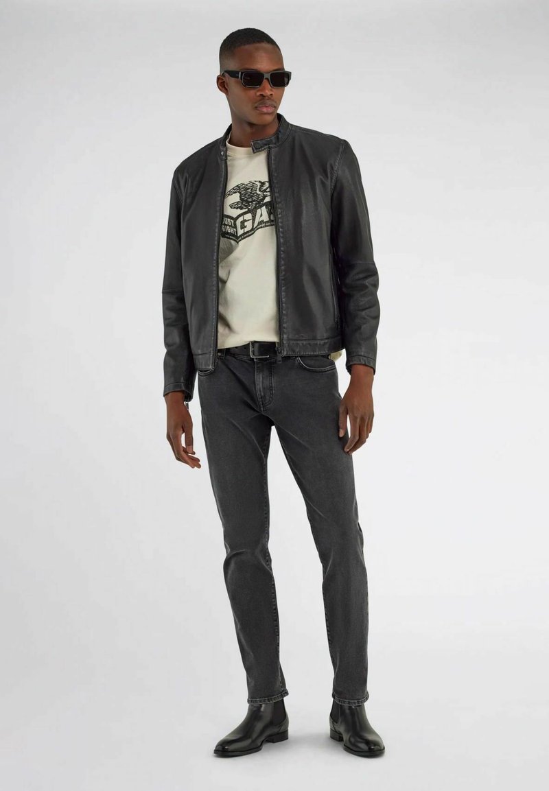 Sort læderjakke, cremefarvet grafisk t-shirt, grå jeans og sorte ankelstøvler. Model iført mørke solbriller, der står imod en ensfarvet baggrund.