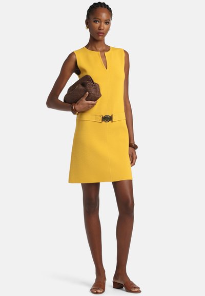 Femeie purtând o rochie galbenă fără mâneci cu curea în față, ținând o poșetă maro împletită și purtând sandale și brățări maro.