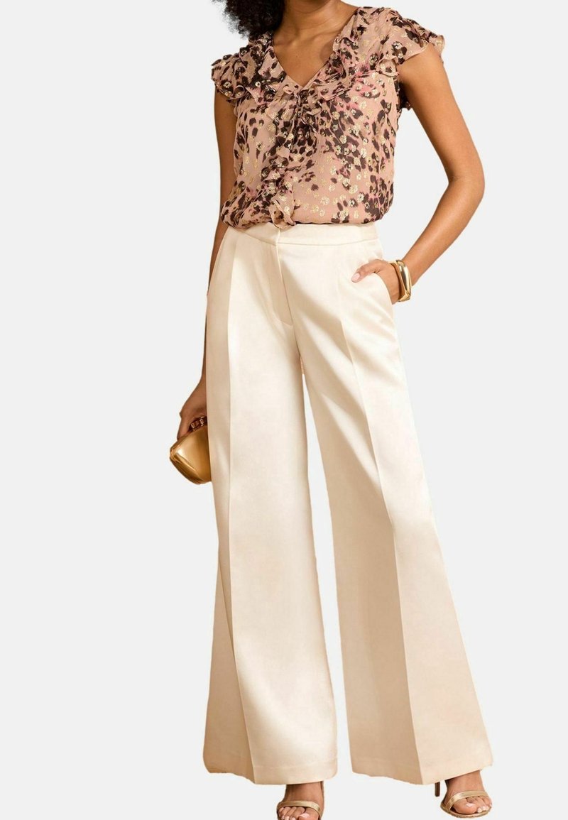 Floral print blouse in light pink and brown, with ruffled sleeves. Cream wide-leg pants with a smooth texture. Gold clutch and sandals.