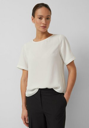 Frau mit zurückgebundenen Haaren, die eine kurzärmelige weiße Bluse und eine schwarze Hose trägt, mit einer Hand in der Tasche stehend vor grauem Hintergrund.