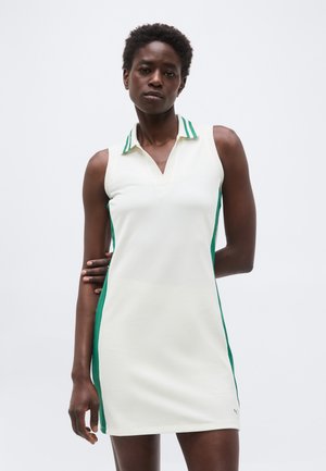 Γυναίκα με κοντά σγουρά μαλλιά που φοράει άσπρο αμάνικο φόρεμα με πράσινες πλευρικές λωρίδες και γιακά με πράσινες ρίγες, στέκεται μπροστά σε απλό φόντο.