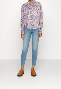 Camicia viola a fantasia floreale con maniche lunghe, abbinata a jeans blu chiaro e mocassini marroni con suola spessa.