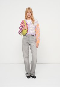 Έγχρωμη κοντή ζακέτα με μοτίβα σε μωβ, πορτοκαλί και ροζ. Γκρι τζιν παντελόνι ψηλόμεσο και μαύρες μπαλαρίνες. Μικρή πράσινη τσάντα με λουρί.