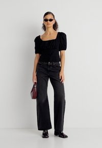 Femme portant un haut noir à manches bouffantes, un jean noir taille haute, des bottes noires, des lunettes de soleil, tenant un petit sac à main rouge foncé, posant devant un fond blanc.