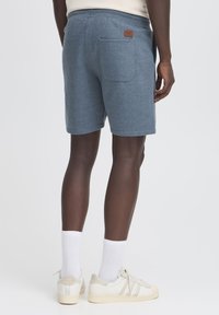 Shorts bleus pour hommes en tissu polaire, dotés d'une poche arrière et d'un patch en cuir marron. Associés à des chaussettes blanches et des baskets crème.