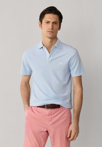 Uomo che indossa una polo a maniche corte azzurro chiaro infilata nei pantaloni rosa con cintura marrone, in piedi contro uno sfondo chiaro uniforme.