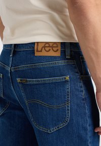 Mörkblå denimjeans med en brun läderetikett som har "Lee" på midjan. Utmärkande sömnad på fickorna. Smal passform.