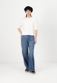 Vit kortärmad topp med avslappnad passform, ihop med vida blå jeans och svarta platta skor. Modellen bär en svart basker.