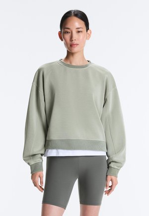 Ženska nosi svetlo zelena prevelika majica s kapuco prek majice bele barve in temno zelenih kolesarskih hlač, stoji pred enobarvno ozadje.