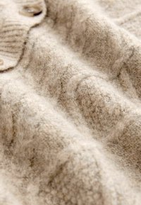 Tekstureeritud beež villane kardigan ribatud krae ja suurte nuppudega, näidates pehmet, umbset pinda ja õrnalt lainetavat kangast.