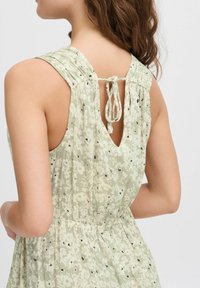 Robe florale vert clair avec un tissu texturé, des plis aux épaules et un système de fermeture à nouer à l'arrière, présentant de petites touches noires sur l'ensemble.