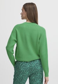 Zeleni rebrasti pulover s okruglim izrezom, raglan rukavima i opuštenim krojem. Uparen s hlačama u uzorcima od menta zelene i crne boje s geometrijskim oblicima.