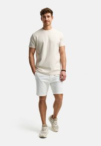 Camiseta corta de manga con un diseño simple en color crema, combinada con pantalones cortos blancos. El modelo lleva zapatillas beige y blancas. Diseño sencillo, textura suave y mínimos acentos.