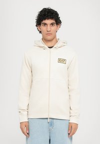 Felpa con zip beige, dotata di cappuccio con cordino, texture morbida e logo EA7 davanti su una targhetta rettangolare dorata. Presenta tasche laterali.