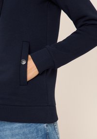 Veste zippée marine en tissu lisse, dotée d'une poche latérale avec un bouton argenté et de poignets et d'un ourlet côtelés.