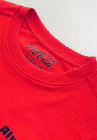 Crvena pamučna majica s okruglim izrezom. Ima crni grafički tekst sprijeda i Coca-Cola logo unutar ovratnika. Glatka tekstura.