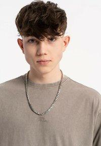 Model w jasnoszarym, krótko rękawowym T-shircie z subtelnym logo i srebrnym naszyjnikiem. Włosy są stylizowane w luźne fale.