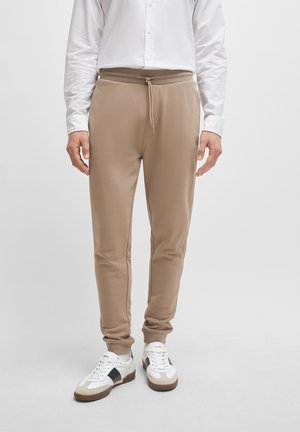 Mann trägt beige Jogginghosen mit Kordelzug an der Taille, weißes Hemd und weiße Turnschuhe mit schwarzen und braunen Details vor einfachem Hintergrund.