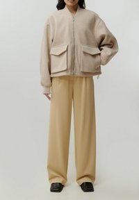 Beige fleece jacket met ritssluiting aan de voorkant, twee grote klepzakken, geribbelde manchetten, gedragen over beige wijde broek en zwarte schoenen.