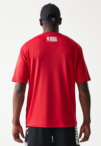 Κόκκινη φανέλα μπάσκετ με ύφασμα δίχτυ, κοντά μανίκια και λογότυπο NBA στην πλάτη. Φοριέται με μαύρο σορτς που φέρει λευκή επωνυμία.