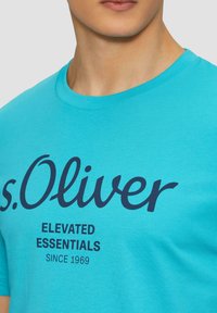 Türkoosist puuvillast T-särk, ümmarguse kaeluse, lühikeste varrukate ja tumedates sinistes trükitud brändinguga, "s.Oliver ELEVATED ESSENTIALS alates 1969."