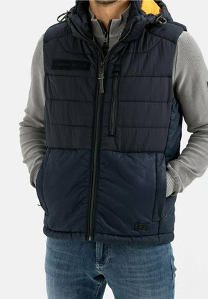 Marineblauer Pufferwesten mit gestepptem Design, abnehmbarer Kapuze in Orange, vorderer Reißverschluss und seitlichen Taschen; getragen über einem grauen Langarmshirt.