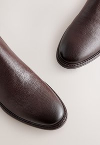 Brune læderankelstøvler med en tekstureret finish, spids tå og kontrasterende glatte detaljer ved tåhætten og sålkanten.