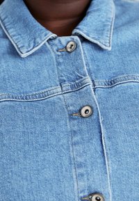 Gaiši zils džinsu jaka ar norādītu apkakli, sudraba pogām un redzamām šuvēm. Iezīmē klasisku piegriezumu un maigu faktūru.