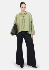 Blusa verde claro con cuello y lazos frontales, combinada con jeans oscuros de pierna ancha. La modelo lleva un bolso marrón oscuro. Textura de tela suave.