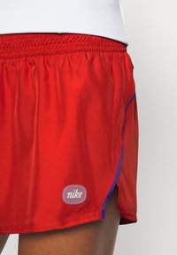 Červené sportovní šortky s elastickým pasem, modrým lemováním a malým logem Nike blízko spodního okraje na stehně osoby.
