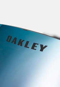 Blauwe getinte zonnebril met een reflecterende lens en de zwarte tekst "OAKLEY" gedrukt op de bovenrand. Strak, modern ontwerp.