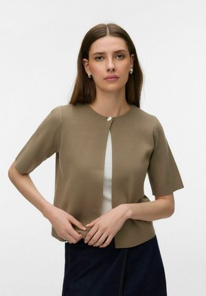 Vrouw met steil bruin haar draagt een olijfgroene kortärmelig cardigan over een wit topje, gecombineerd met een donkere rok, staand tegen een effen achtergrond.