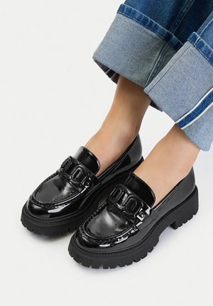 Mocasines negros gruesos y brillantes con detalle decorativo de hebilla, usados con vaqueros azules remangados y pies descalzos sobre un fondo liso.