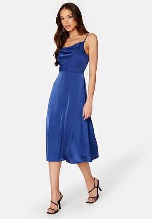 Blauwe satijnen midi-jurk met een plooikraag, dunne bandjes en een vloeiende rok. Gefabriceerd met zwarte sandalen met hoge hakken en bandjes.