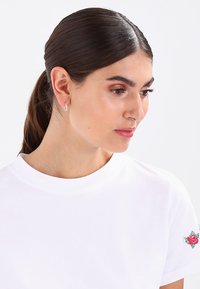 Camiseta blanca de algodón con cuello redondo y bordado floral en rojo y verde en el hombro izquierdo, con una textura suave.