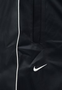 Fekete széldzseki fehér csíkozással és fehér Nike logóval. Könnyű, texturált anyagból készült, cipzáras zsebfelülettel.