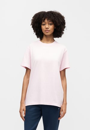 Γυναίκα με σγουρά μαλλιά, που φοράει ένα ανοιχτό ροζ κοντομάνικο μπλουζάκι και σκούρο μπλε παντελόνι, στέκεται μπροστά σε ένα απλό φωτεινό φόντο.