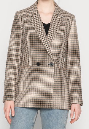Personne portant un blazer pied-de-poule marron et noir avec des boutons noirs, sur un haut noir et un jean bleu clair, debout devant un fond uni.