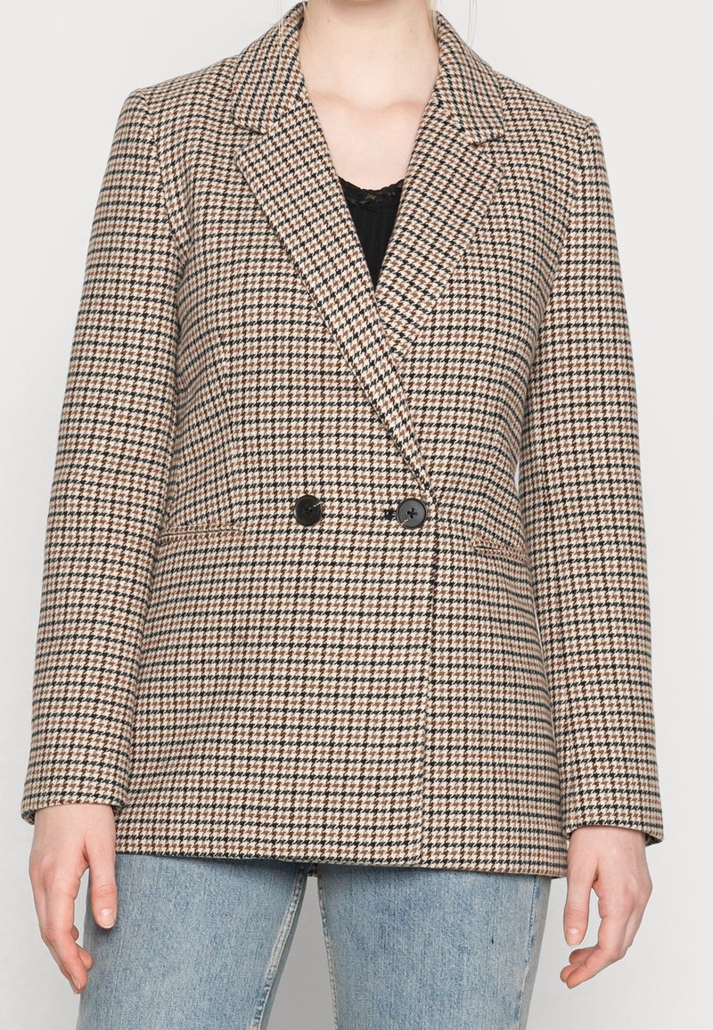 Personne portant un blazer pied-de-poule marron et noir avec des boutons noirs, sur un haut noir et un jean bleu clair, debout devant un fond uni.