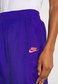 Fialové lehké šortky s elastickým pasem a bočními kapsami, s růžovým logem Nike na levé nohavici. Hladká textura.