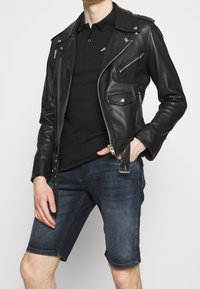 Černá kožená motorkářská bunda se stříbrnými zipy a patentkami, nošená přes černé polo tričko. V kombinaci s tmavými džínovými šortkami a sponou na opasek.