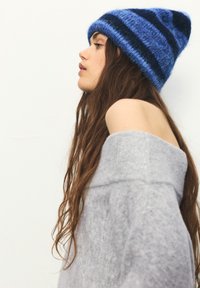 Blau gestreifte Mütze aus weichem, flauschigem Material; grauer schulterfreier Pullover mit strukturierter Oberfläche; langes, welliges Haar sichtbar.