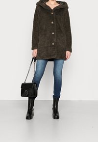 Olivengrøn, fuzzy frakke med hætte, der har knaplukning og to lommer. Parret med blå jeans og sorte hæle støvler, mens der holdes en sort taske.