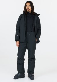 Zwarte skijas en -broek gemaakt van duurzaam materiaal; voorzien van ritsen, zakken en een gestructureerde afwerking. G gedragen met zwarte skistuks.
